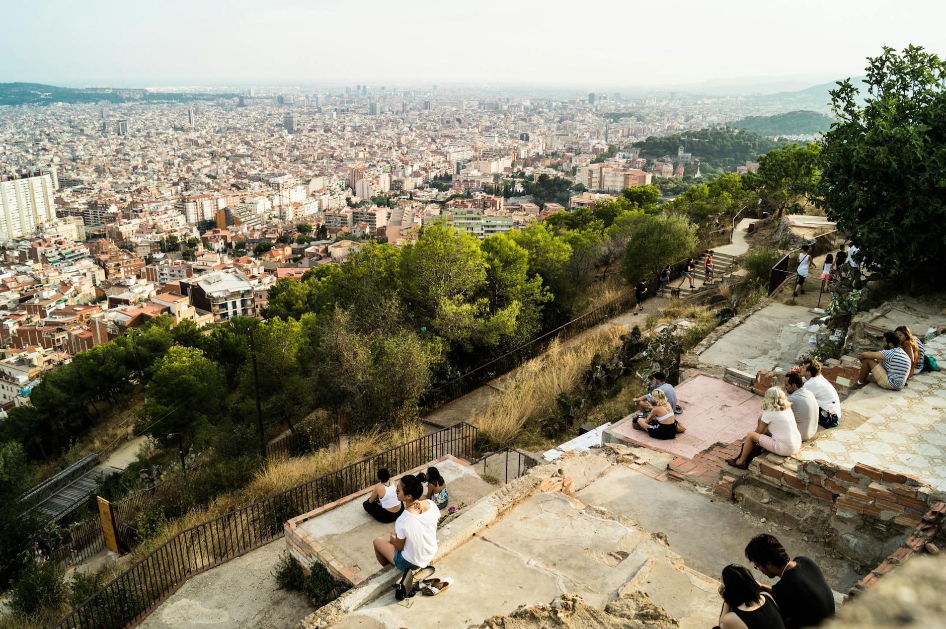 A panoramic view of Barcelona from the hills, showcasing the cityscape with people sitting and enjoying the scenery among greenery.