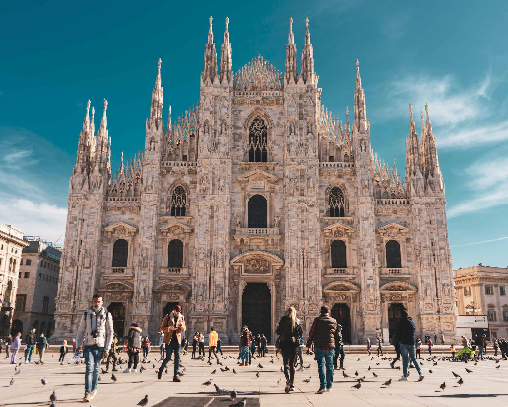The Duomo di Milano, a stunning Gothic cathedral, with visitors walking in front and pigeons scattered on the ground in Piazza del Duomo.