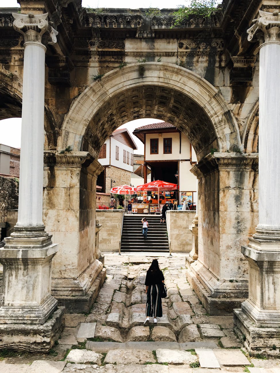 "Timeless Passage – A Woman Walks Through Hadrian’s Gate in Antalya" A woman walking through Hadrian's Gate, an ancient marble archway, with Ottoman-style buildings and a small market in the background.
