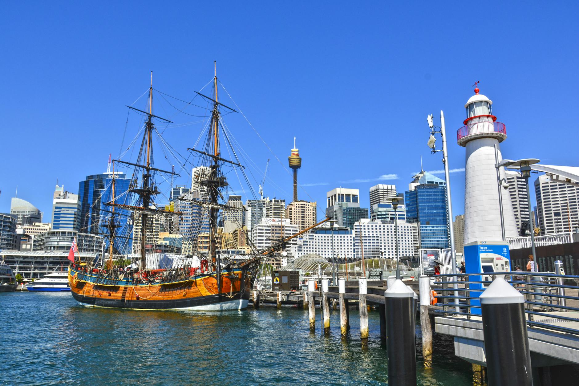 A historic tall ship docked at a marina with the Sydney skyline in the background, featuring modern skyscrapers and a lighthouse.