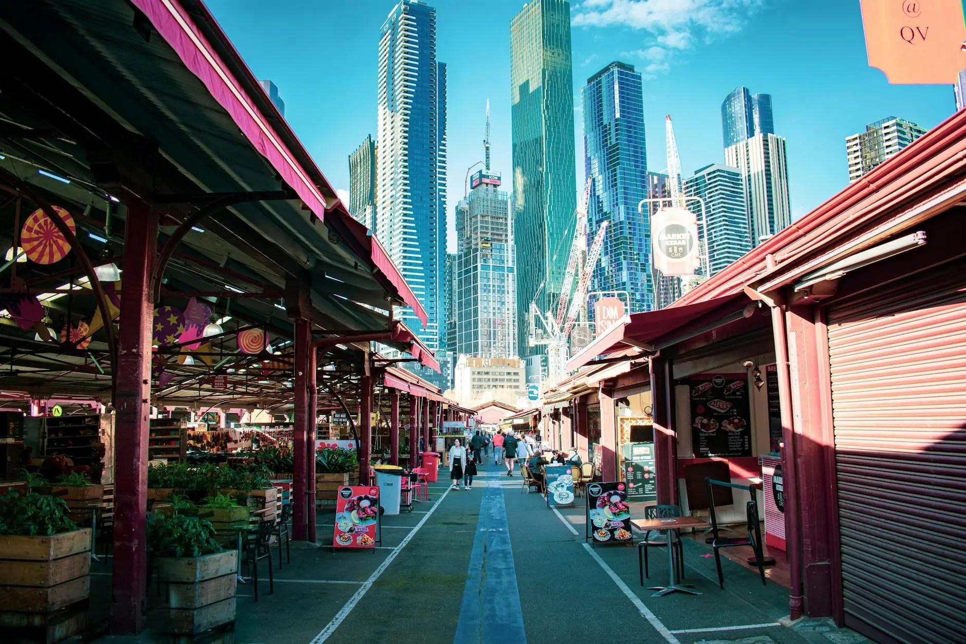 A vibrant view of the Queen Victoria Market in Melbourne, showcasing colorful stalls and a backdrop of tall city skyscrapers.