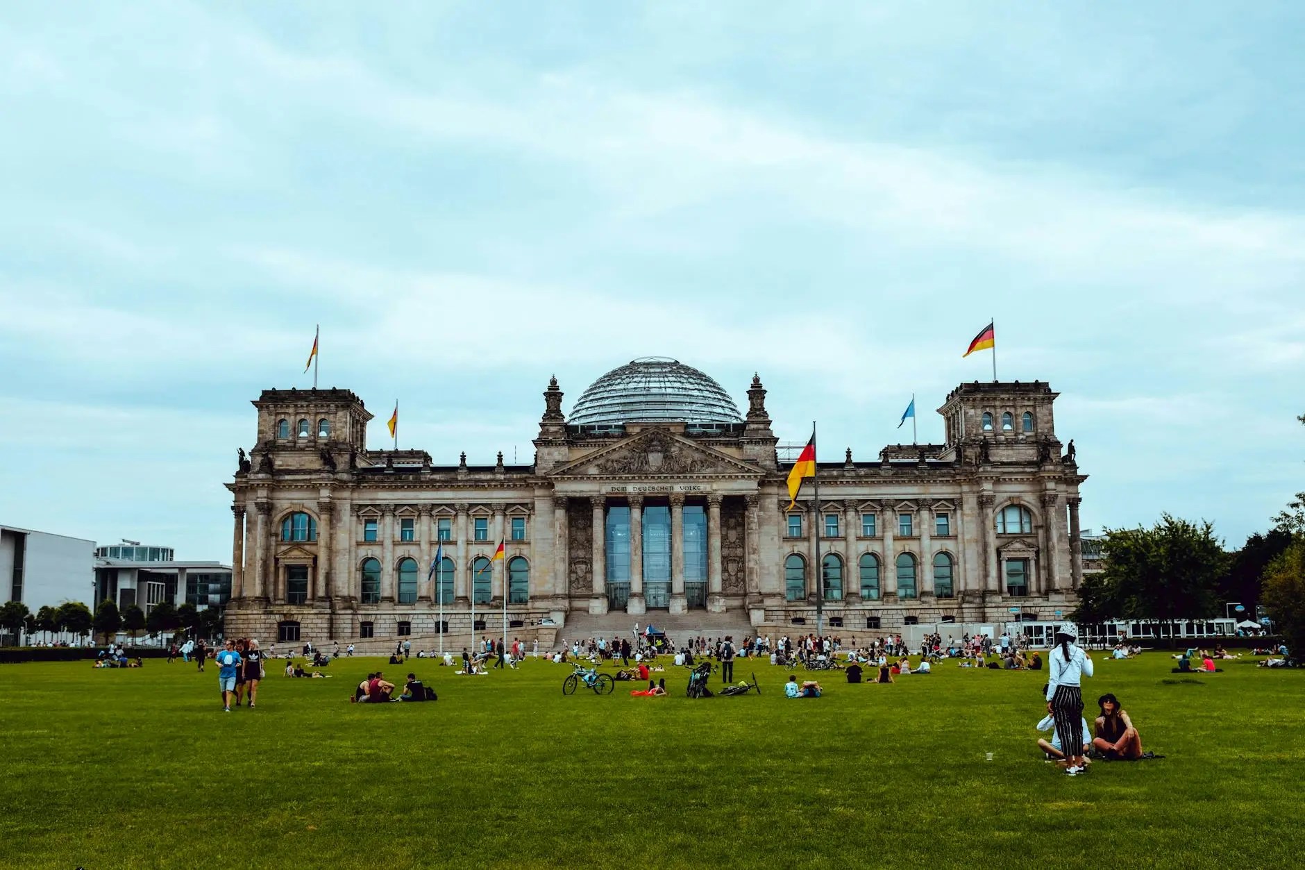 The Reichstag Building in Berlin, featuring its glass dome and surrounded by a large grassy area filled with people enjoying the outdoors.