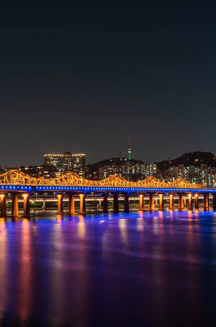 Night view of a brightly lit bridge over a river with a city skyline in the background, showcasing vibrant colors reflecting on the water.
