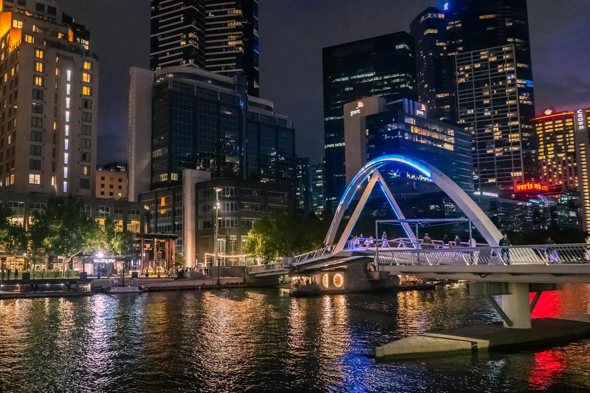 Night view of the Yarra River in Melbourne, showcasing illuminated buildings and a pedestrian bridge.