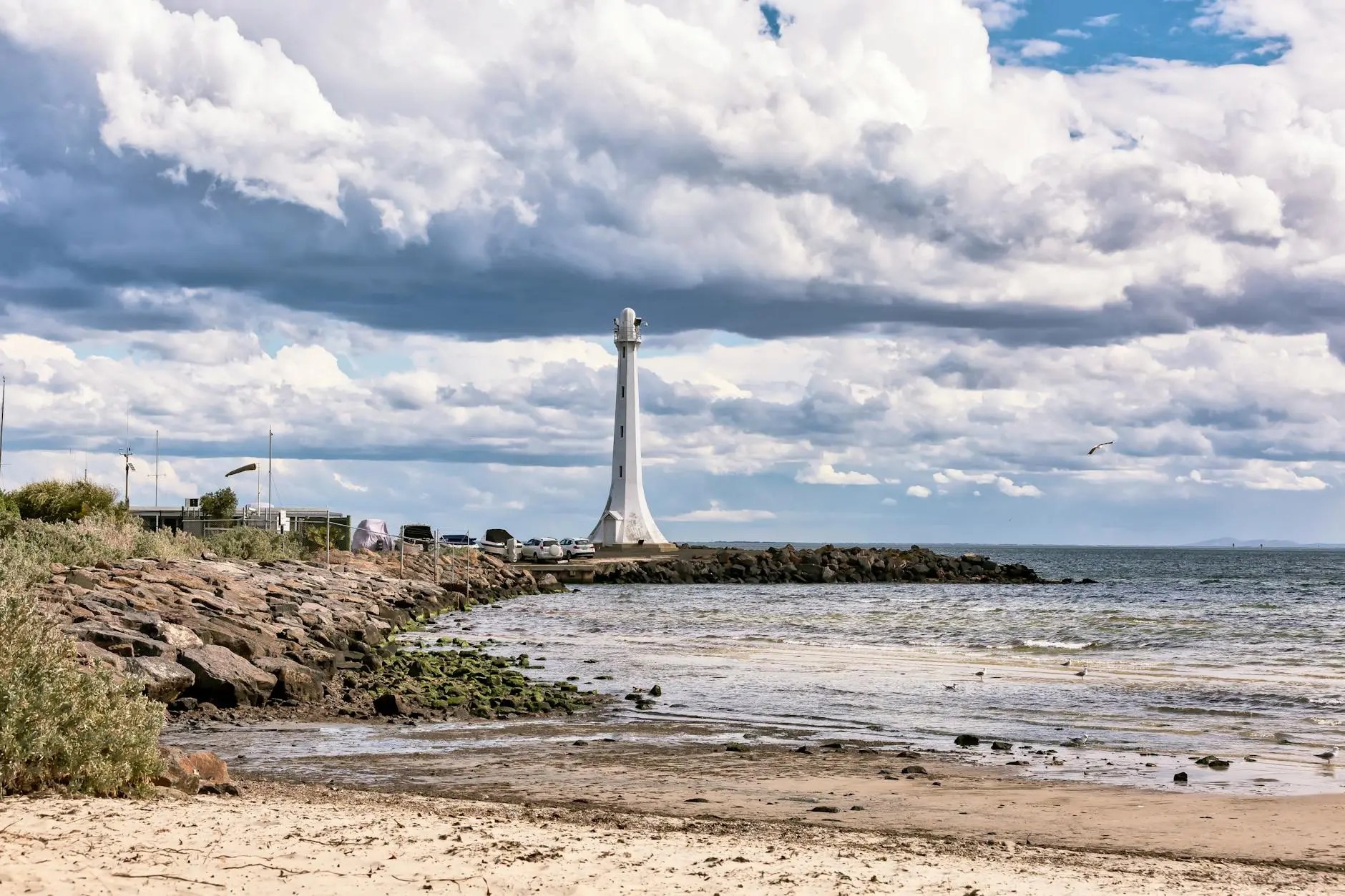 A scenic view of a sandy beach with a lighthouse in the background, featuring a rocky coastline and cloudy sky.