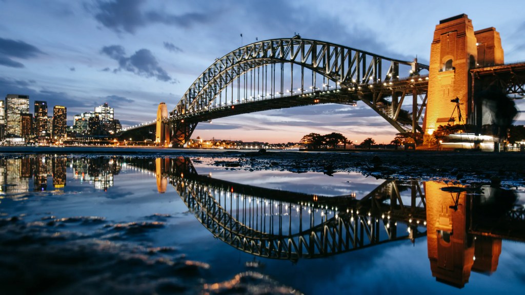 Sydney Harbour Bridge at dusk reflecting in the water, with city skyline in the background.