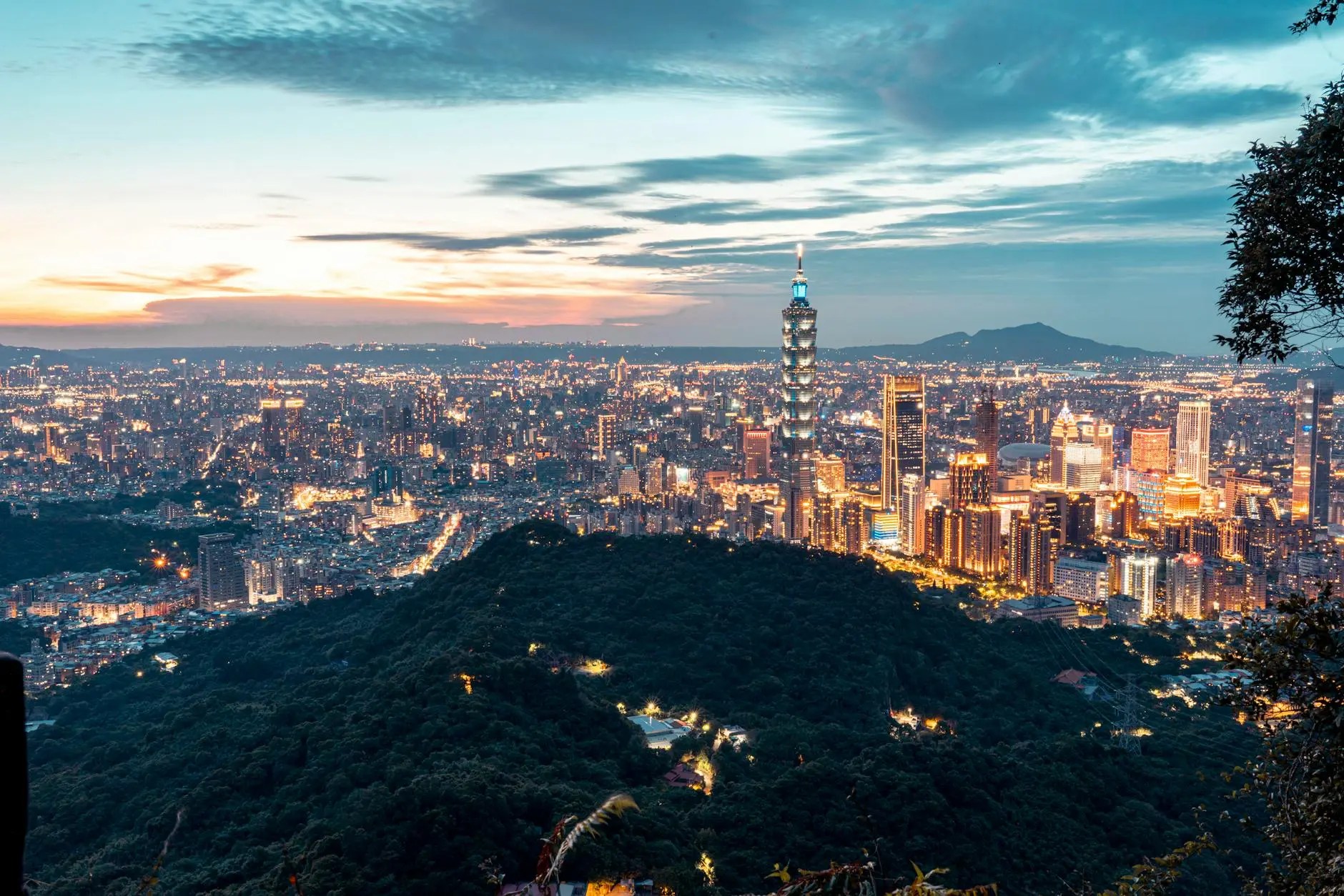 A panoramic view of Taipei at sunset, showcasing the Taipei 101 skyscraper and the city's illuminated skyline amidst rolling hills.