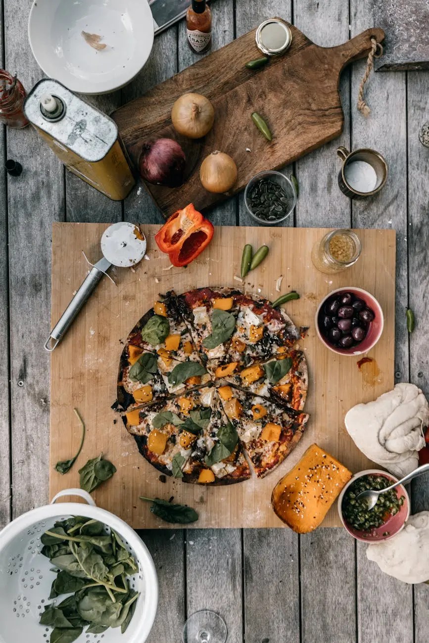 A wooden cutting board with a sliced vegetable pizza topped with spinach and yellow bell peppers, surrounded by various ingredients like onions, a red bell pepper, and spices.