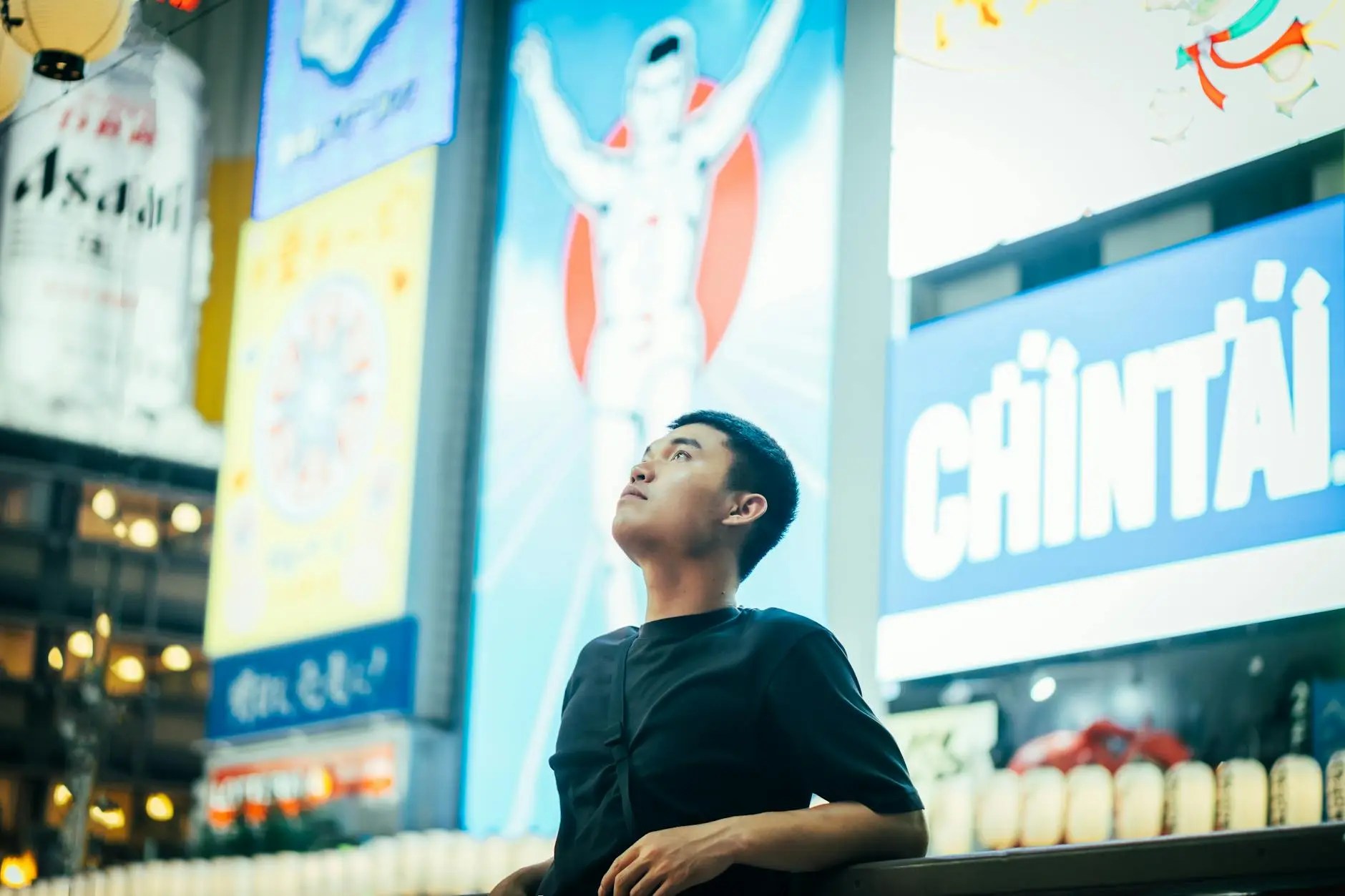 A person gazing up at neon lights and advertisements in Dotonbori, Osaka, surrounded by vibrant city scenery.
