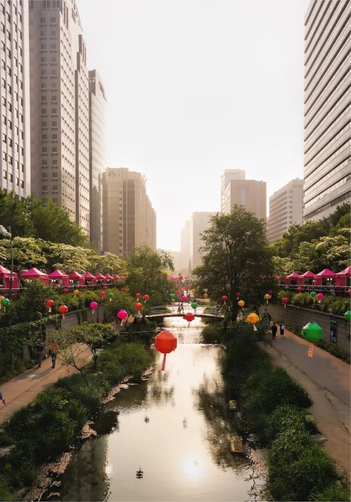 A view of a serene waterway in Seoul lined with colorful lanterns, framed by modern skyscrapers and lush greenery.