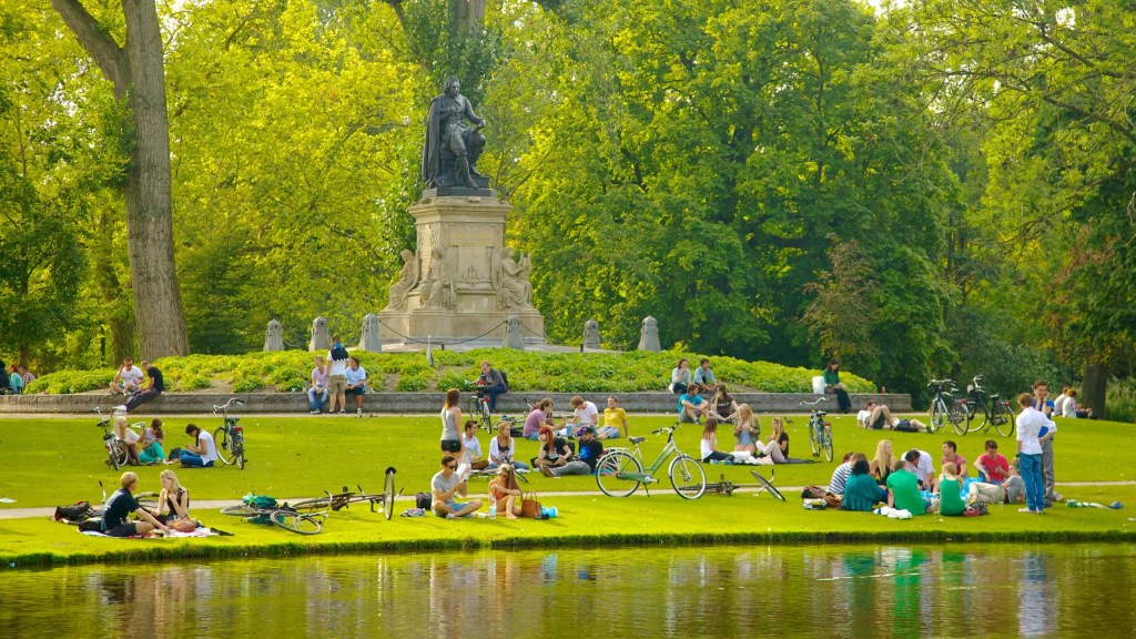 People enjoying a sunny day in Vondelpark, Amsterdam, with bicycles nearby and a statue in the background, surrounded by lush greenery.