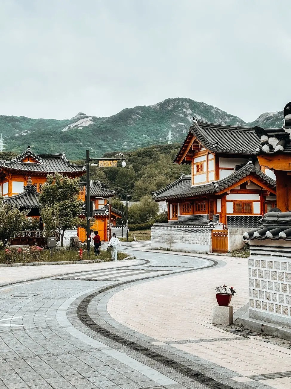 A winding pathway through Bukchon Hanok Village featuring traditional Korean houses surrounded by lush greenery and distant mountains.