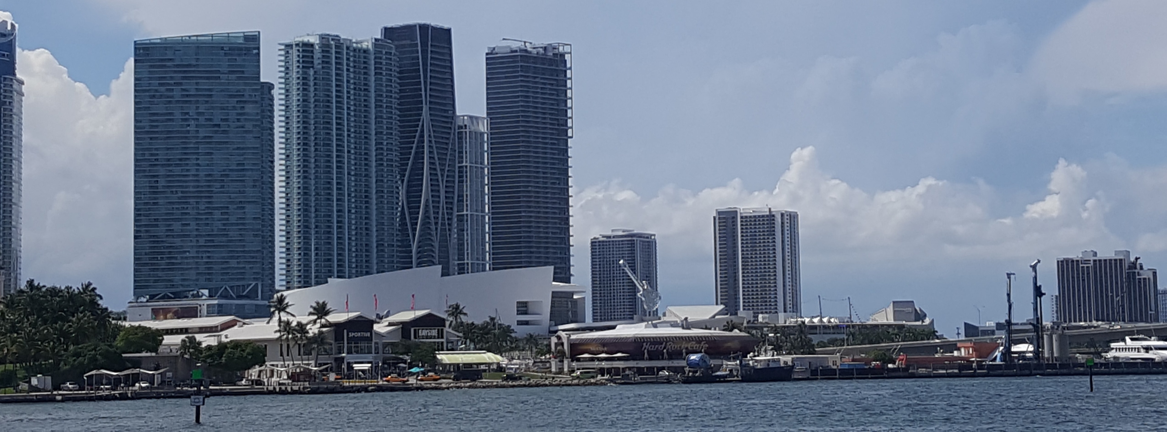 View of Miami's skyline featuring modern skyscrapers and the Hard Rock Cafe, with the waterfront in the foreground and a partly cloudy sky.