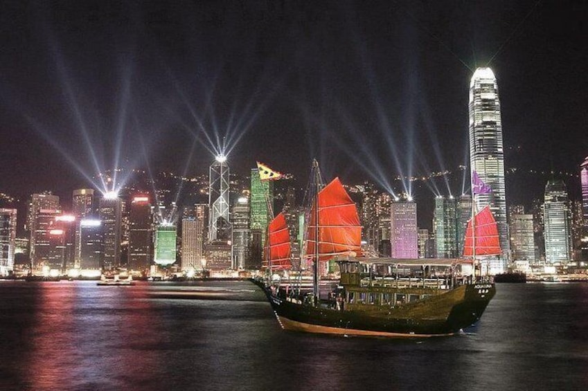 A traditional Chinese junk boat with red sails sailing in Victoria Harbour, Hong Kong, illuminated by vibrant city lights and laser beams from nearby skyscrapers at night.