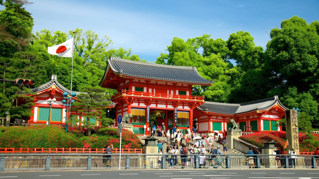 A vibrant, traditional Japanese shrine with a distinctive red and white facade surrounded by lush greenery, featuring visitors exploring the entrance and a Japanese flag waving. Yasaka Shrine