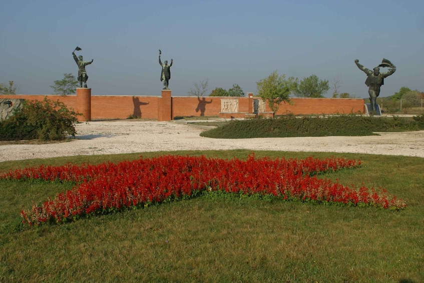 A park featuring a symbolic red star flowerbed in front of statues representing historical figures, set against a clear sky.