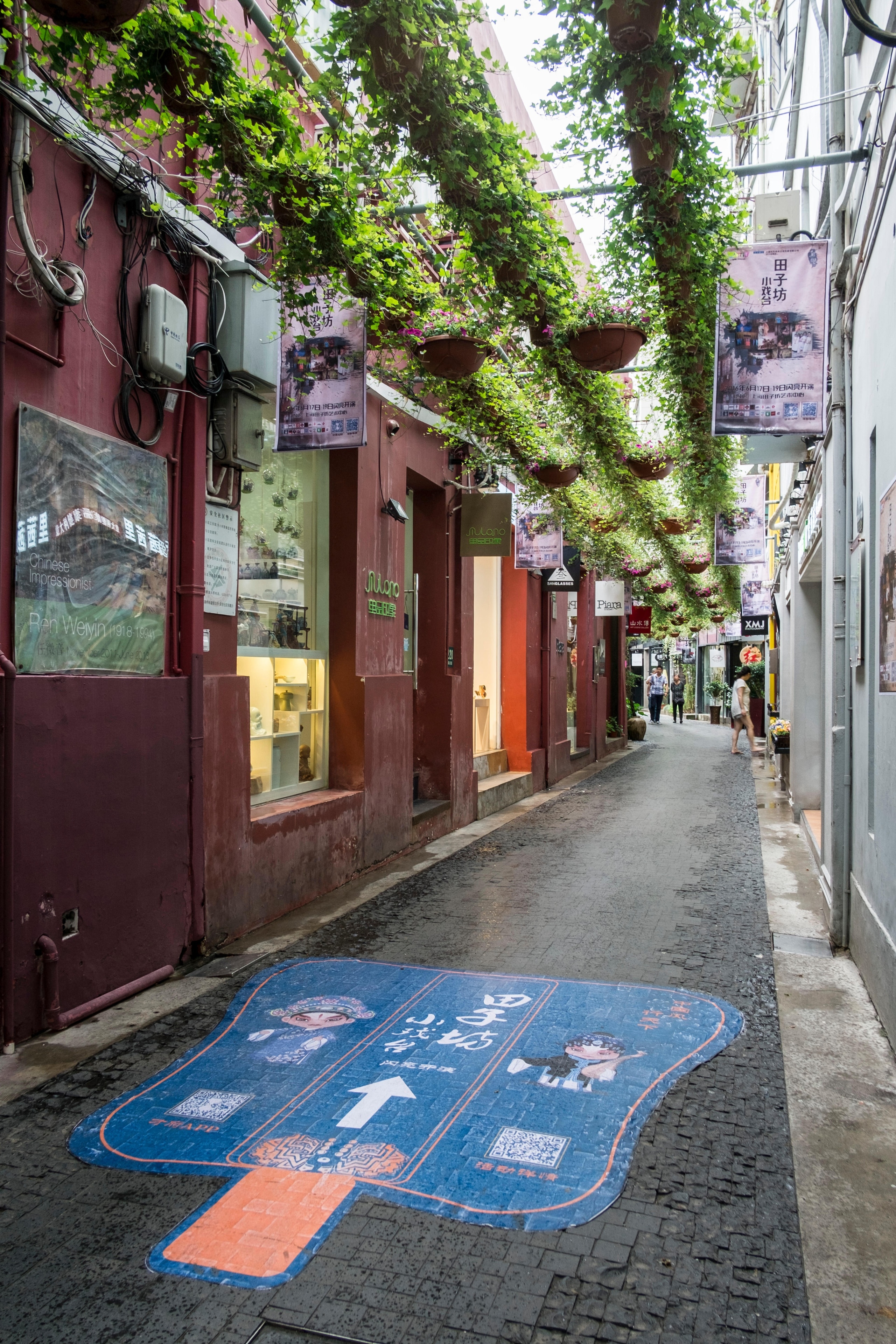 A narrow alley in Shanghai lined with red buildings, adorned with hanging plants and posters, leading to shops and cafes.