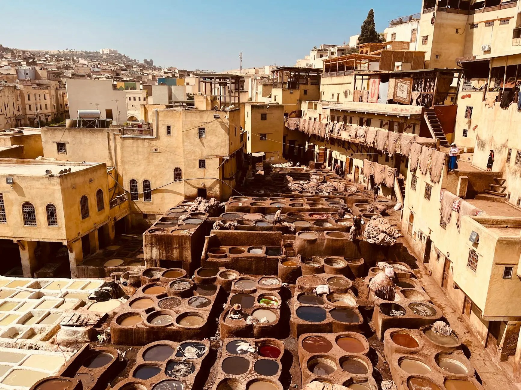 View of traditional Moroccan tanneries in Fez, featuring large dyeing pits surrounded by beige buildings and residents working with leather.