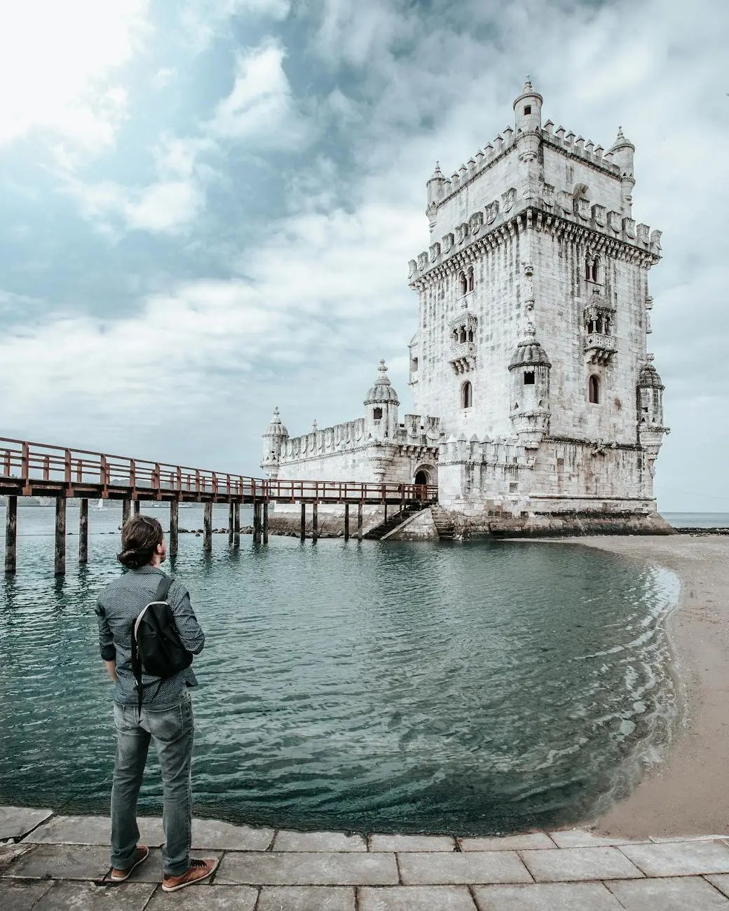 A person standing by the water, gazing at the Belém Tower in Lisbon under a partly cloudy sky.