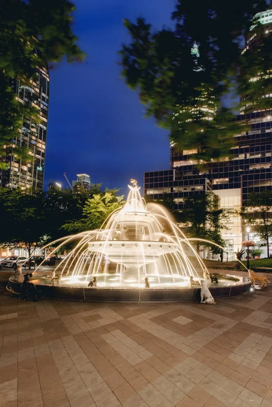 A beautifully lit fountain at night, surrounded by trees and modern buildings in an urban setting.