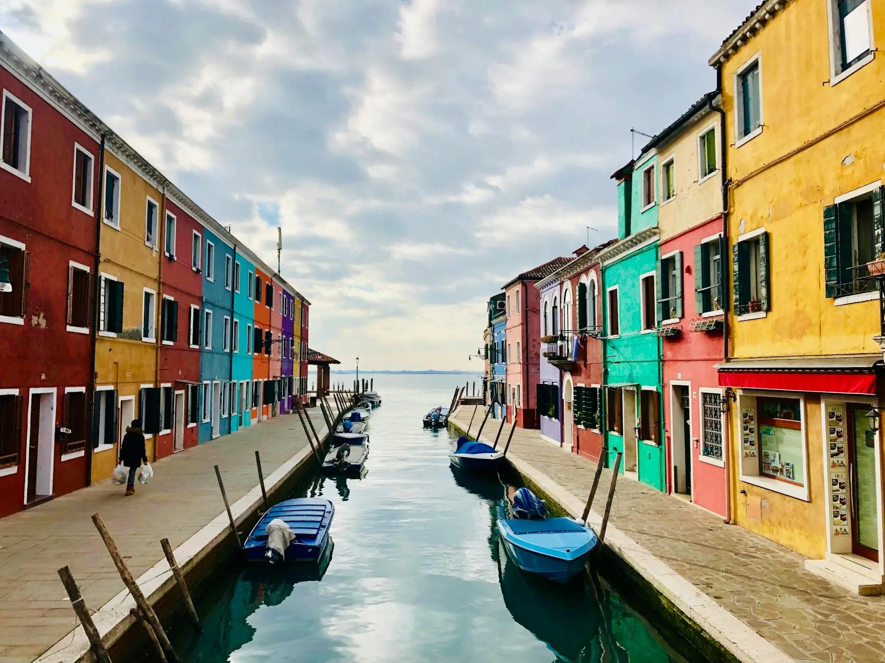 A picturesque canal in Burano, Venice, lined with colorful houses and small boats moored along the waterfront, under a cloudy sky.