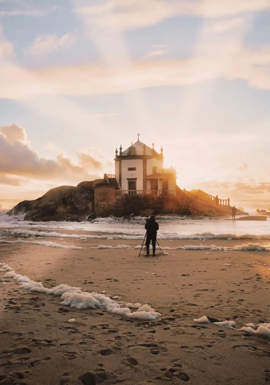 A person capturing a photo on the beach at sunset, with the Capela do Senhor da Pedra visible on a rock in the background.