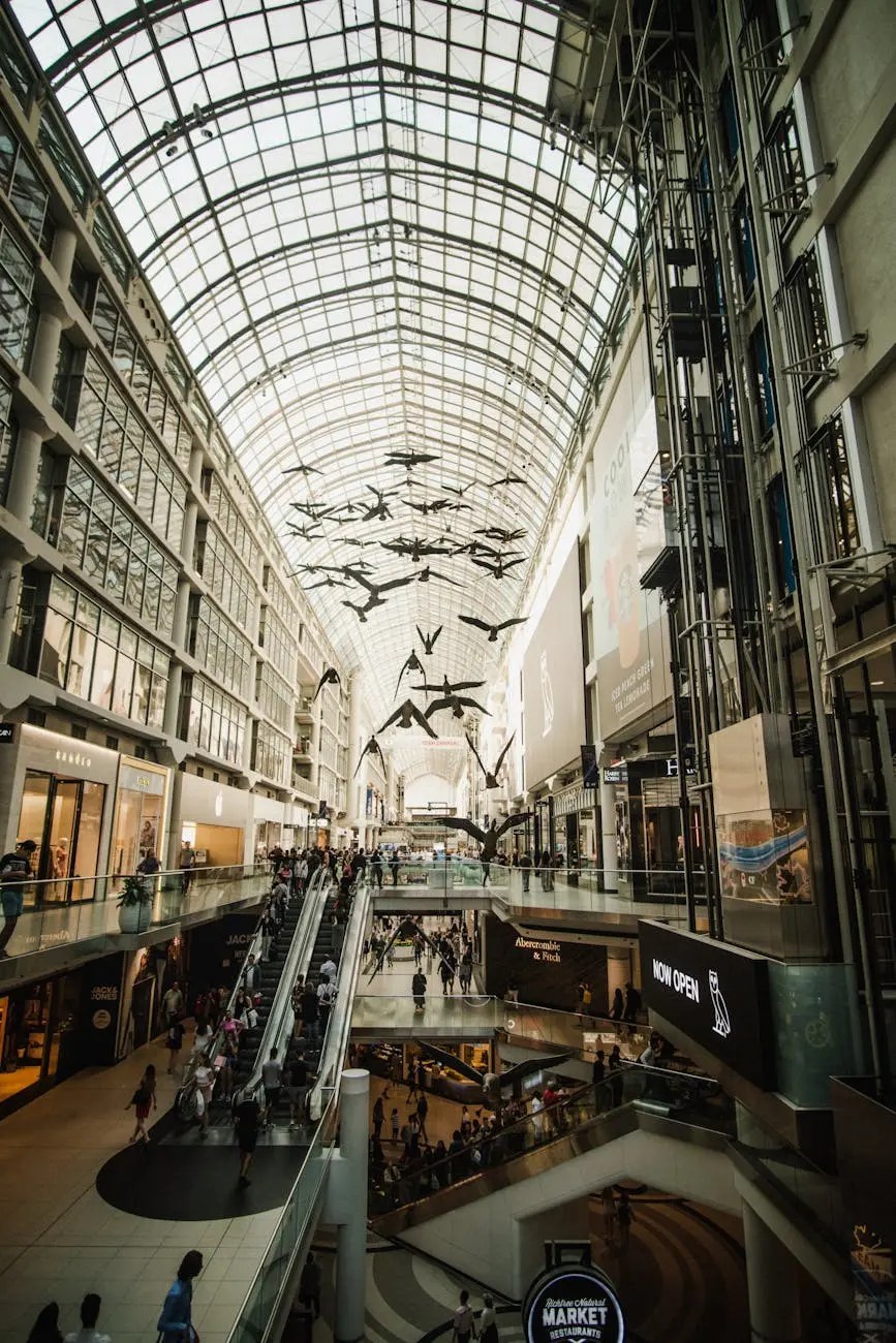 A busy shopping mall with a glass ceiling, featuring escalators and a large flock of birds hanging from the ceiling. Shoppers and visitors are seen walking throughout the space.