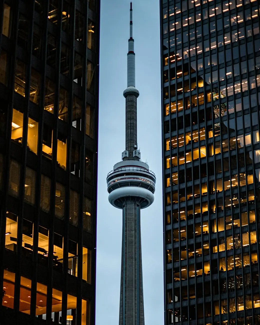 The CN Tower stands tall between two urban buildings, illuminated by warm lights against a twilight sky.