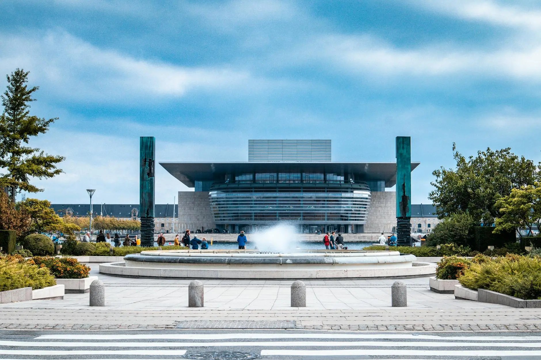 View of the Copenhagen Opera House with a fountain in the foreground, surrounded by greenery and people enjoying the outdoor space.