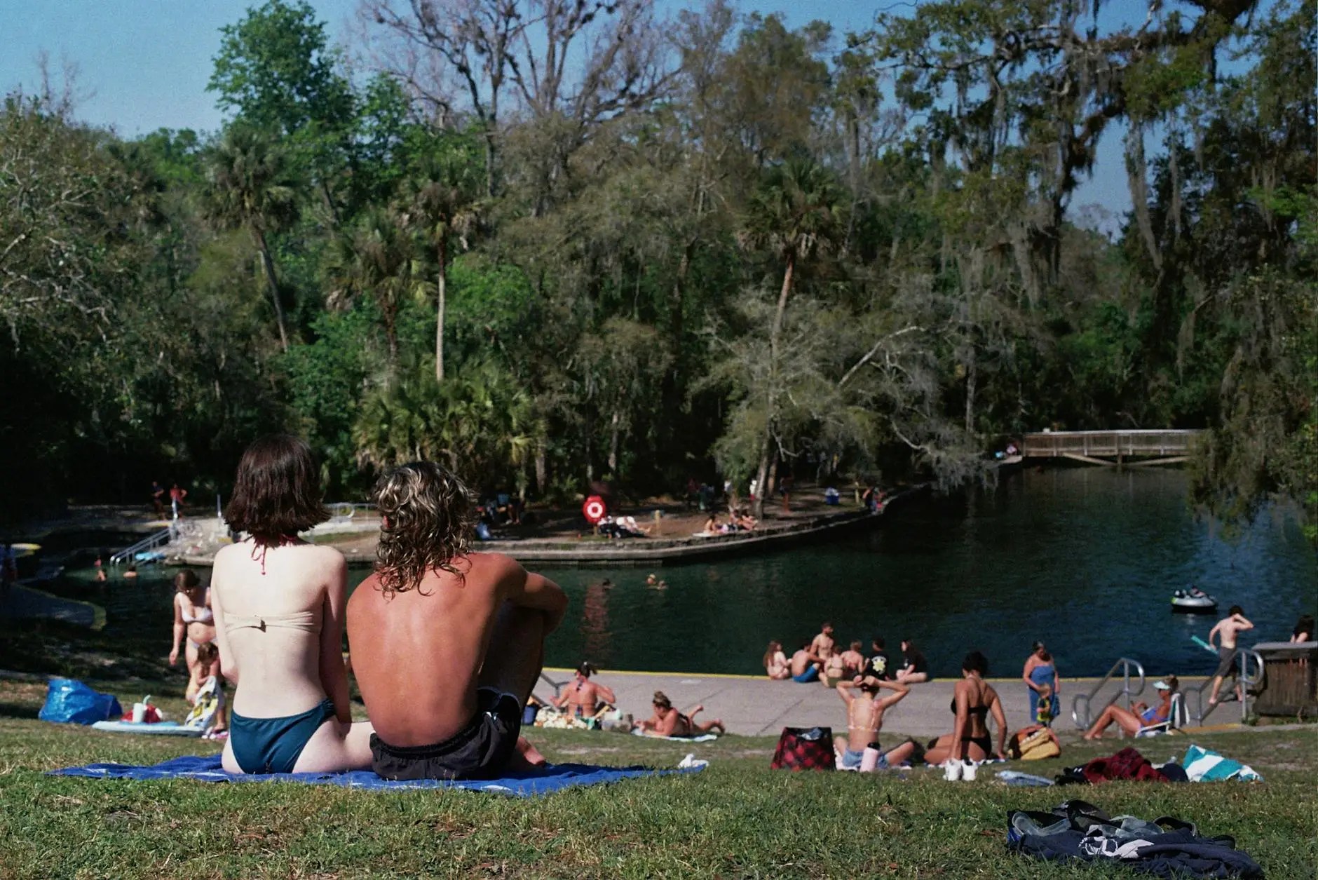 A couple sits on a blanket by a lake, enjoying a sunny day in Florida. In the background, people are swimming and relaxing, surrounded by lush greenery and trees.