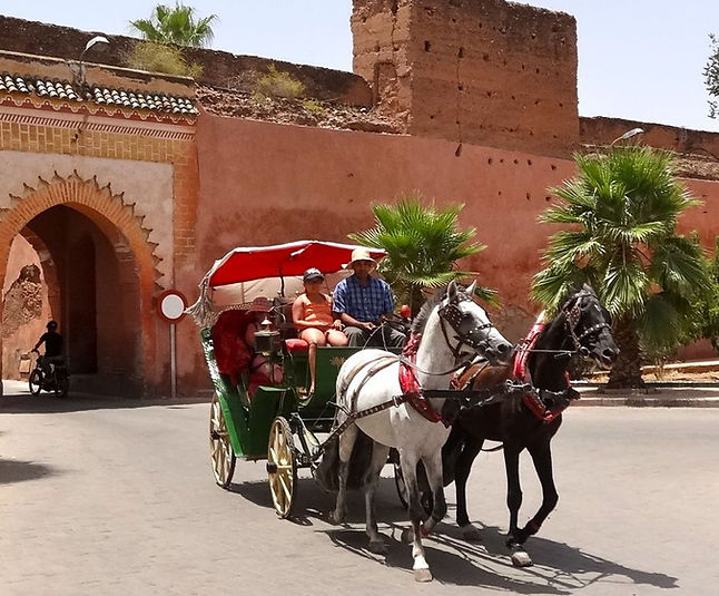 A horse-drawn carriage, known as a caleche, rides through the streets of Marrakech, with passengers seated under a red canopy near historic walls.