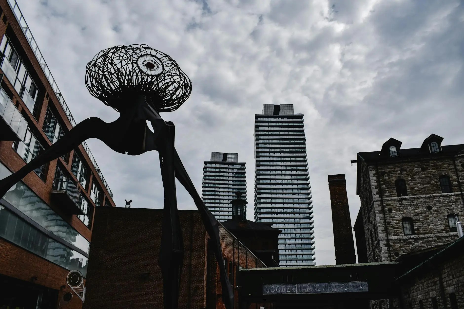 A large sculpture resembling an abstract creature with a spherical head made of wire, set against a backdrop of modern high-rise buildings and a cloudy sky in the Distillery District of Toronto.