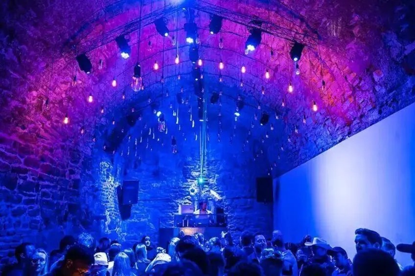 A vibrant nightlife scene in a stone-arched venue, illuminated by colorful lights with a crowd enjoying the atmosphere.