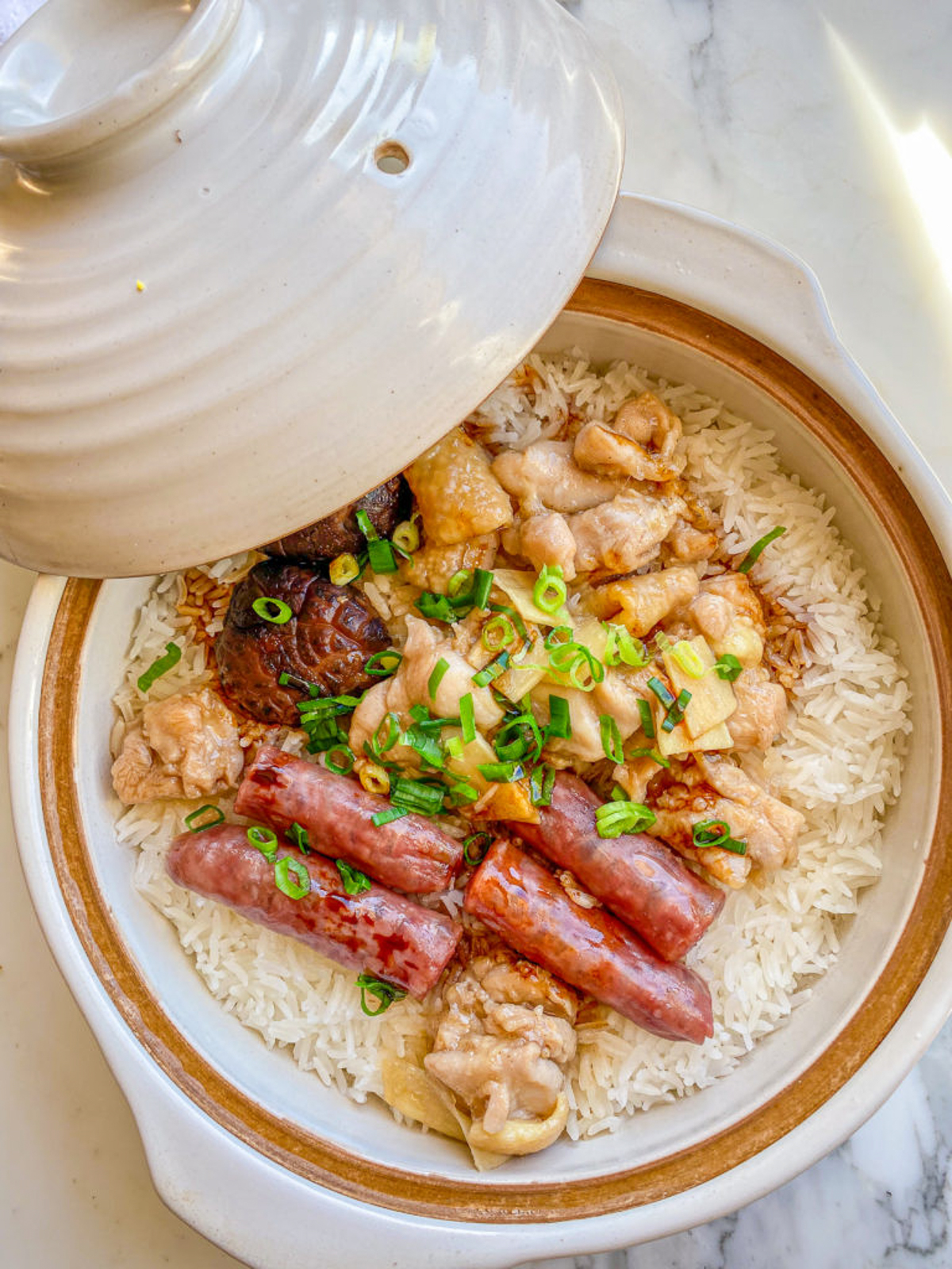 A clay pot filled with rice topped with pieces of chicken, sausages, and garnished with chopped green onions.
