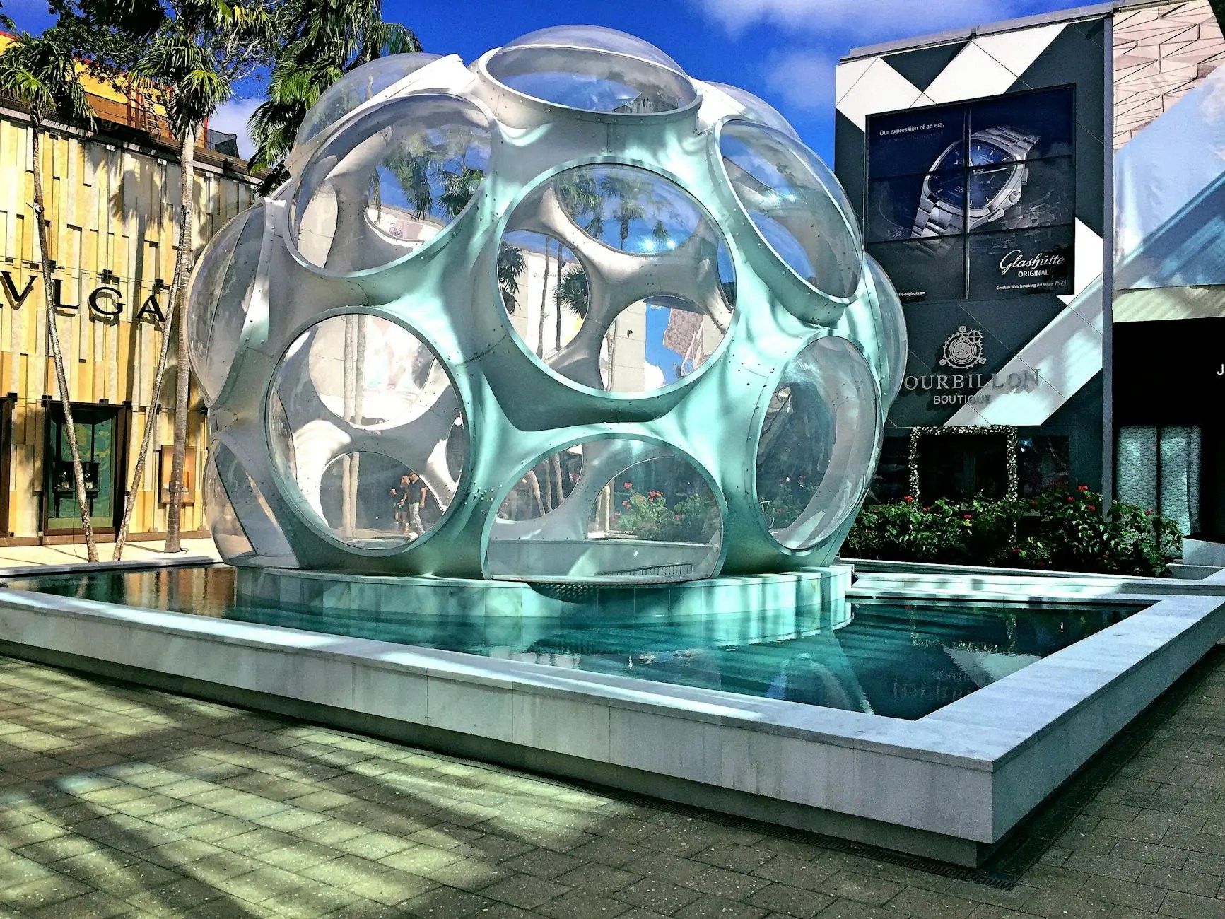 A modern art installation resembling a translucent sphere with circular openings, surrounded by a reflective pool and vibrant greenery, set against a backdrop of stylish boutiques.