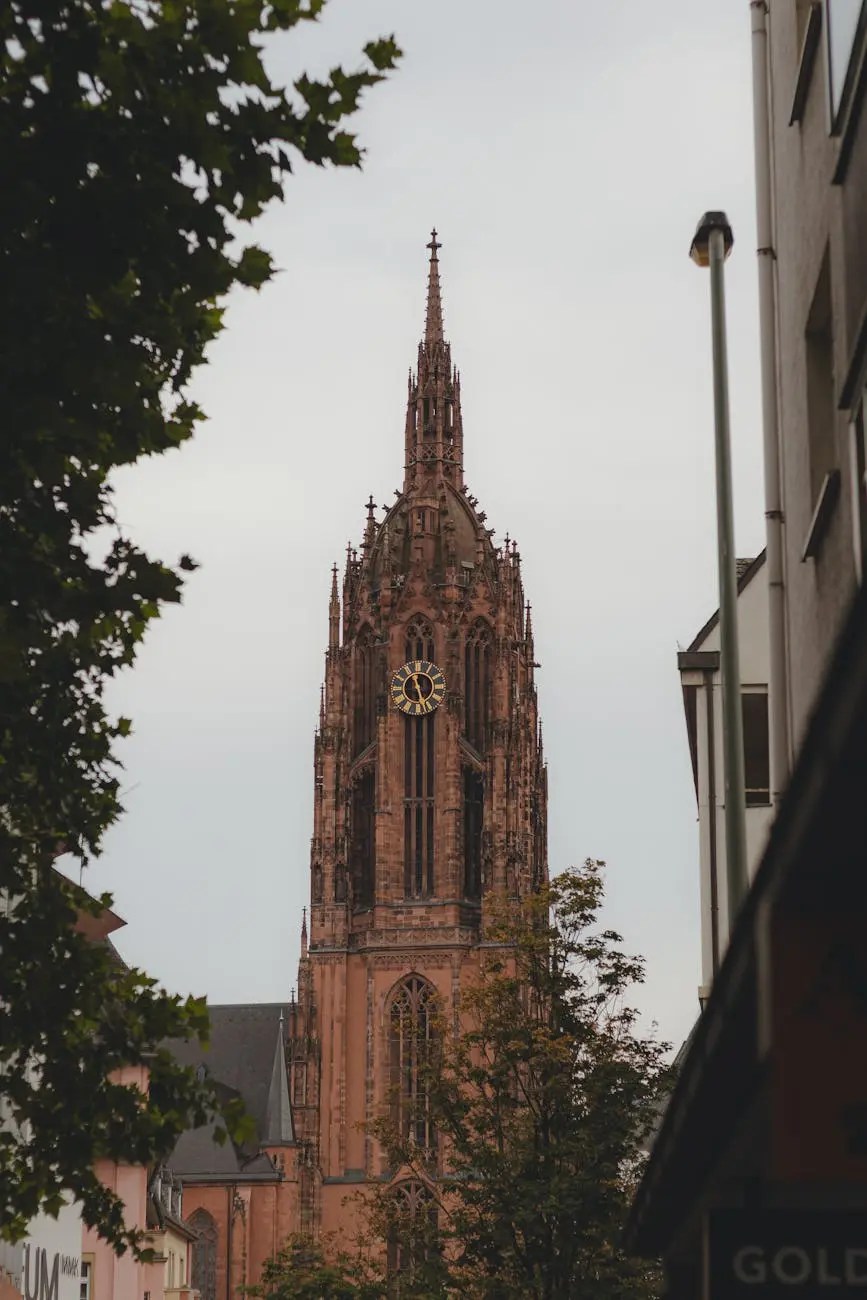 A tall Gothic church tower peeks through a narrow street, surrounded by trees and modern buildings, under a cloudy sky.