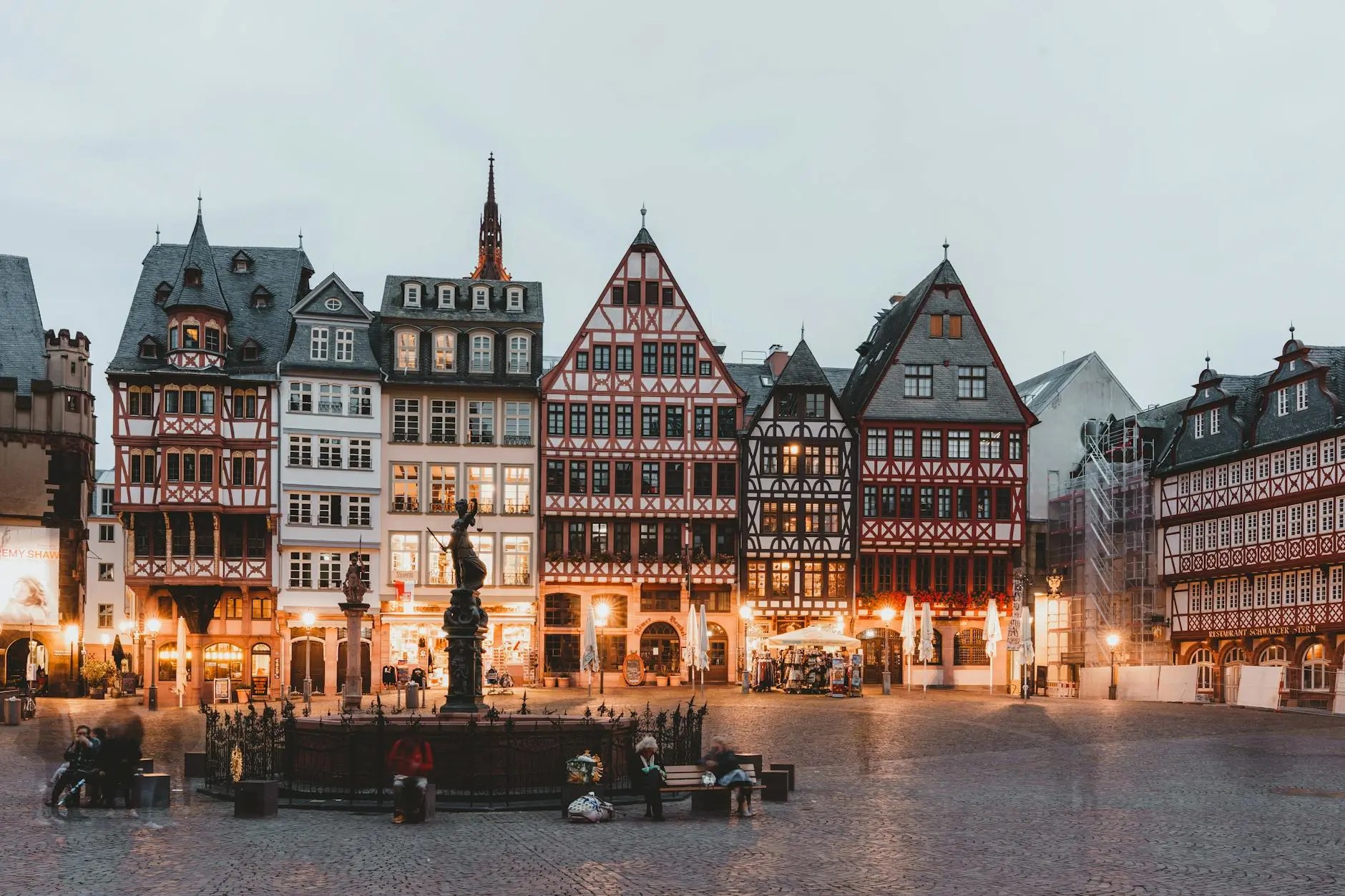 A picturesque view of Römerberg square in Frankfurt, featuring charming half-timbered houses and a statue in the center, illuminated by warm lights during twilight.