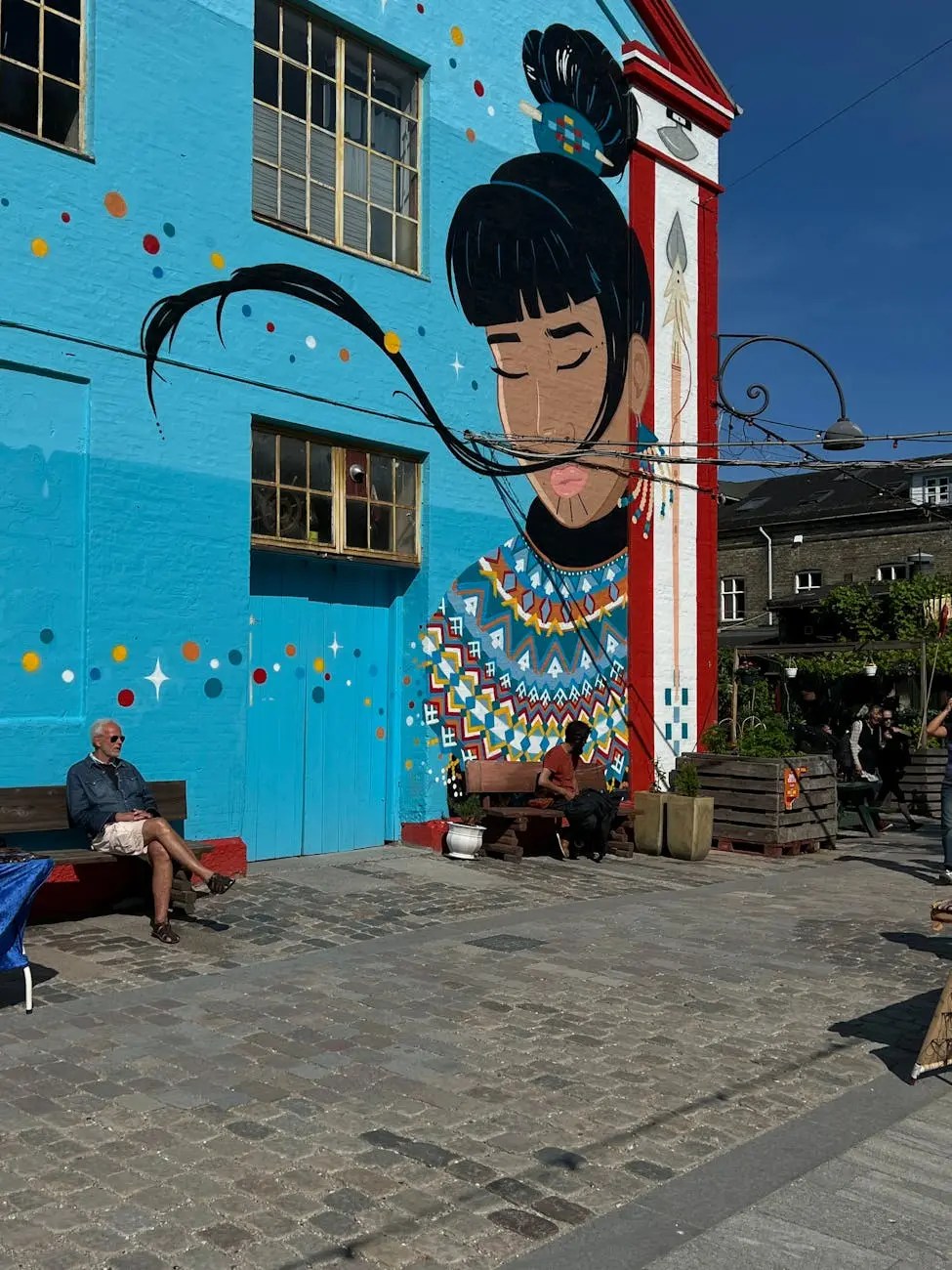 A vibrant mural featuring a woman with long hair against a bright blue wall, with colorful geometric patterns and shapes surrounding her. The scene includes people sitting on benches in front of the mural, with a cobblestone pathway.