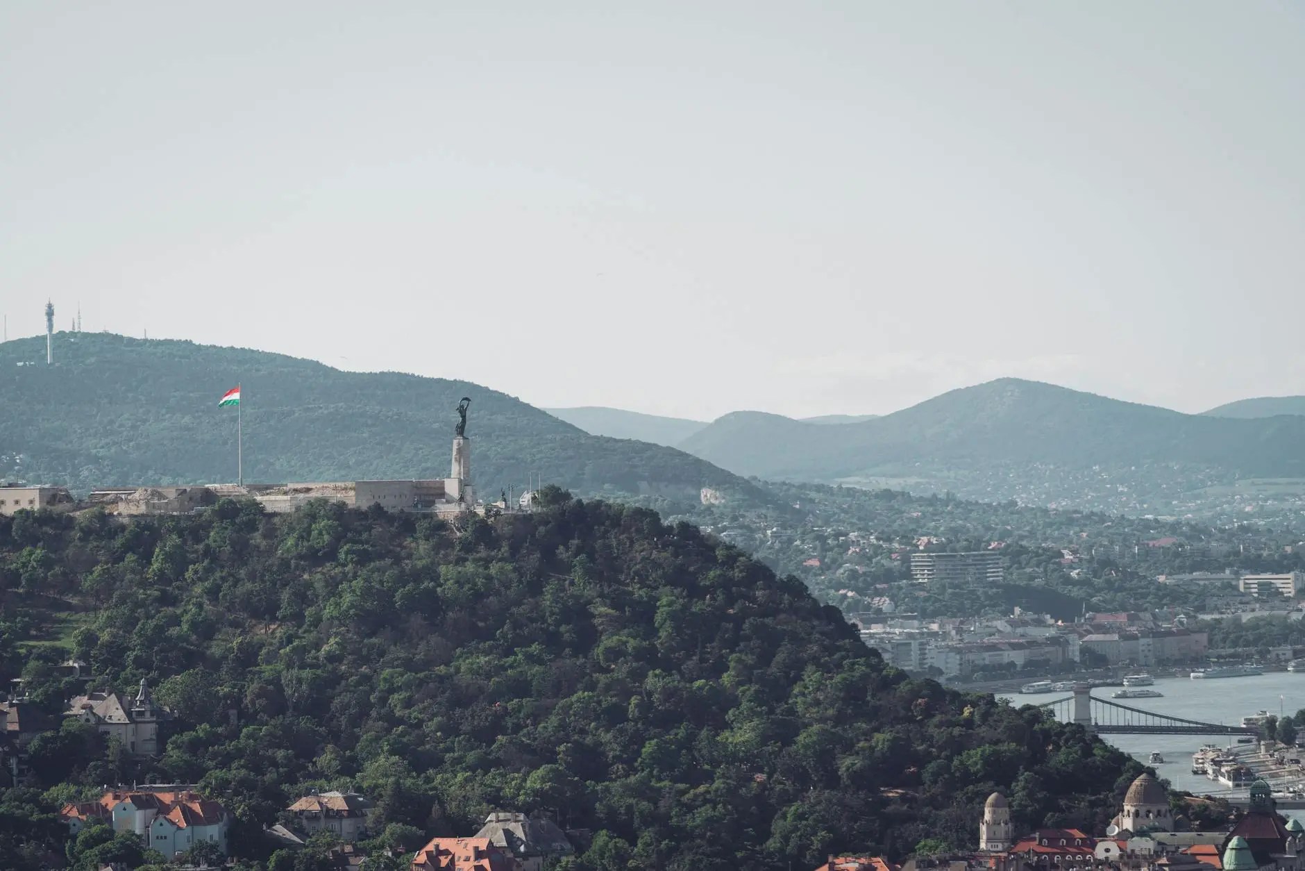 A panoramic view of the Buda Hills in Budapest, featuring a statue on a hilltop and the Danube River in the foreground, with a flag waving in the wind.