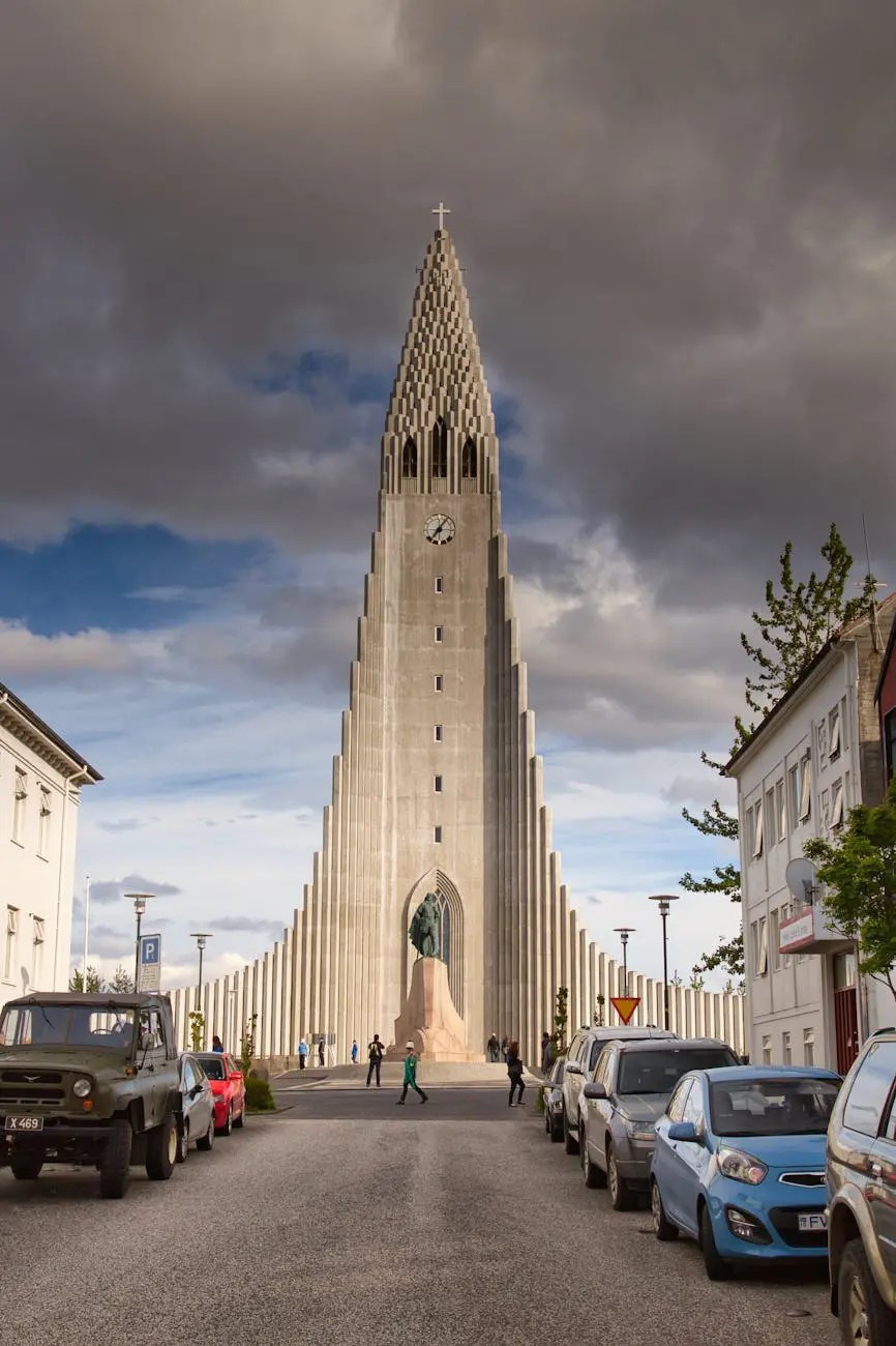A wide-angle view of Hallgrímskirkja, a prominent church in Reykjavik, Iceland, featuring its distinctive tall tower and unique architectural design. Cars are parked along the street, and people are seen walking in front of the church under a partly cloudy sky.