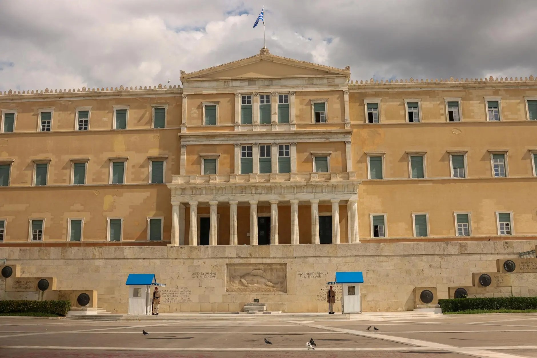 Front view of the Hellenic Parliament building in Athens, featuring classical architecture with columns, a flag flying above, and guards at the entrance.