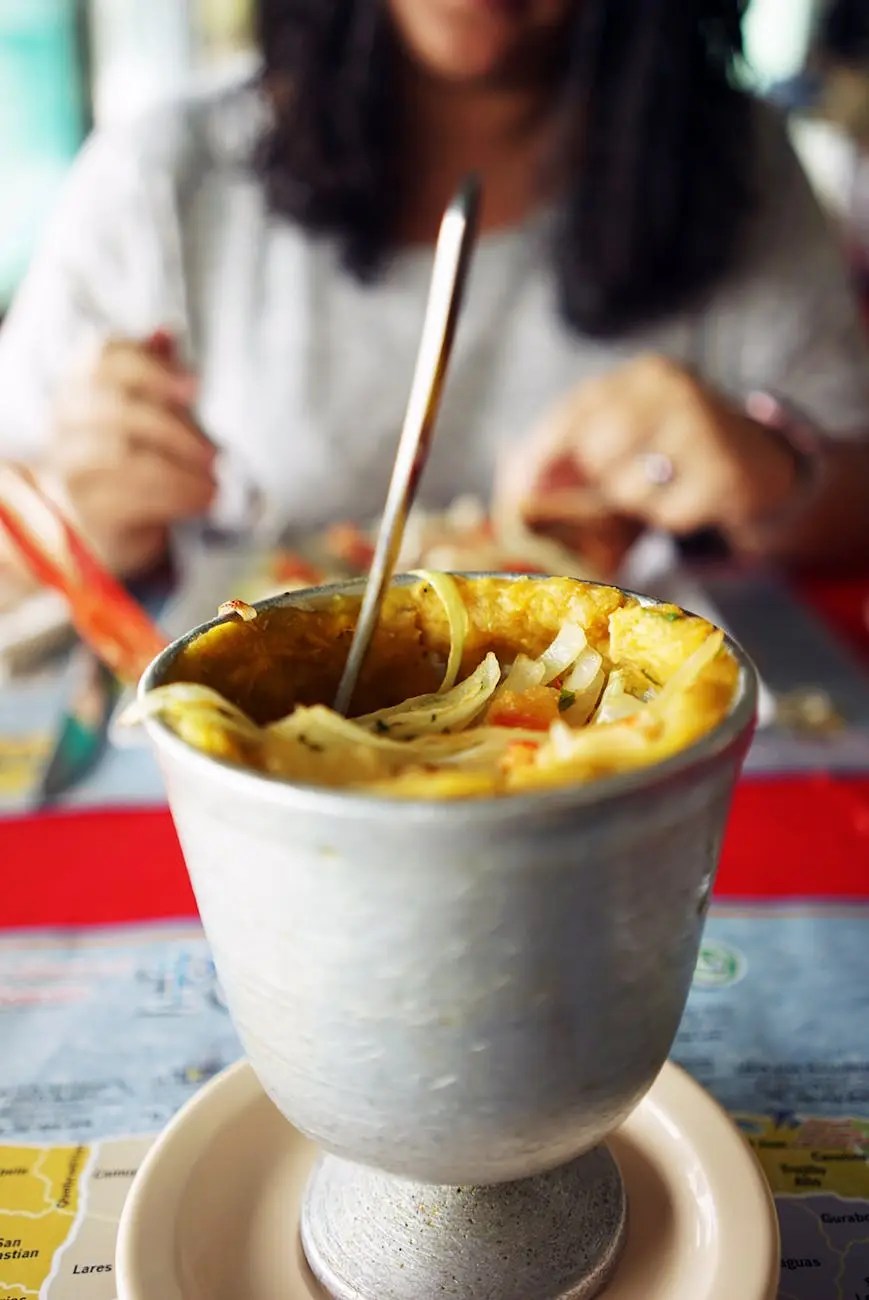 Close-up of a bowl of macaroni soup with a spoon, with a person eating in the background.