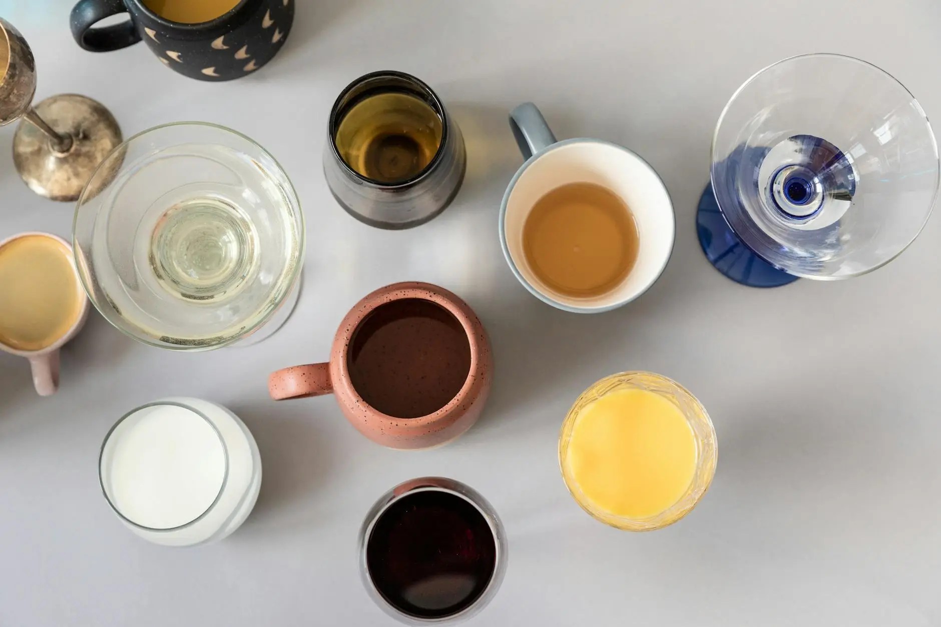 An overhead view of various beverage cups and glasses, including a glass of water, a mug with tea, a clay pitcher, and a wine glass, arranged artistically on a flat surface.