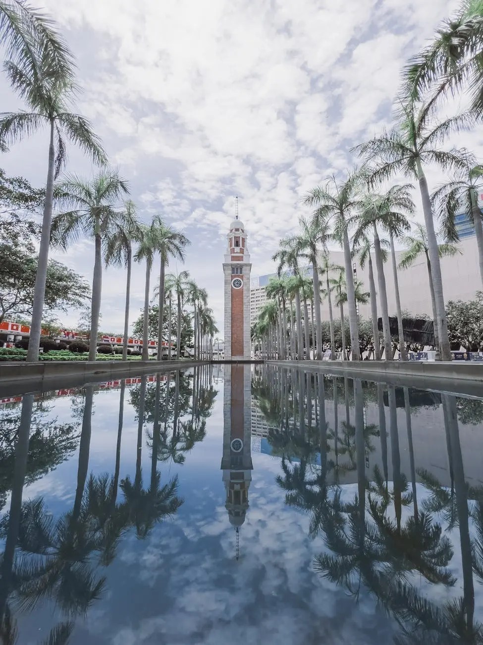 View of a clock tower surrounded by palm trees with a reflection in a still pond under a cloudy sky in Hong Kong.