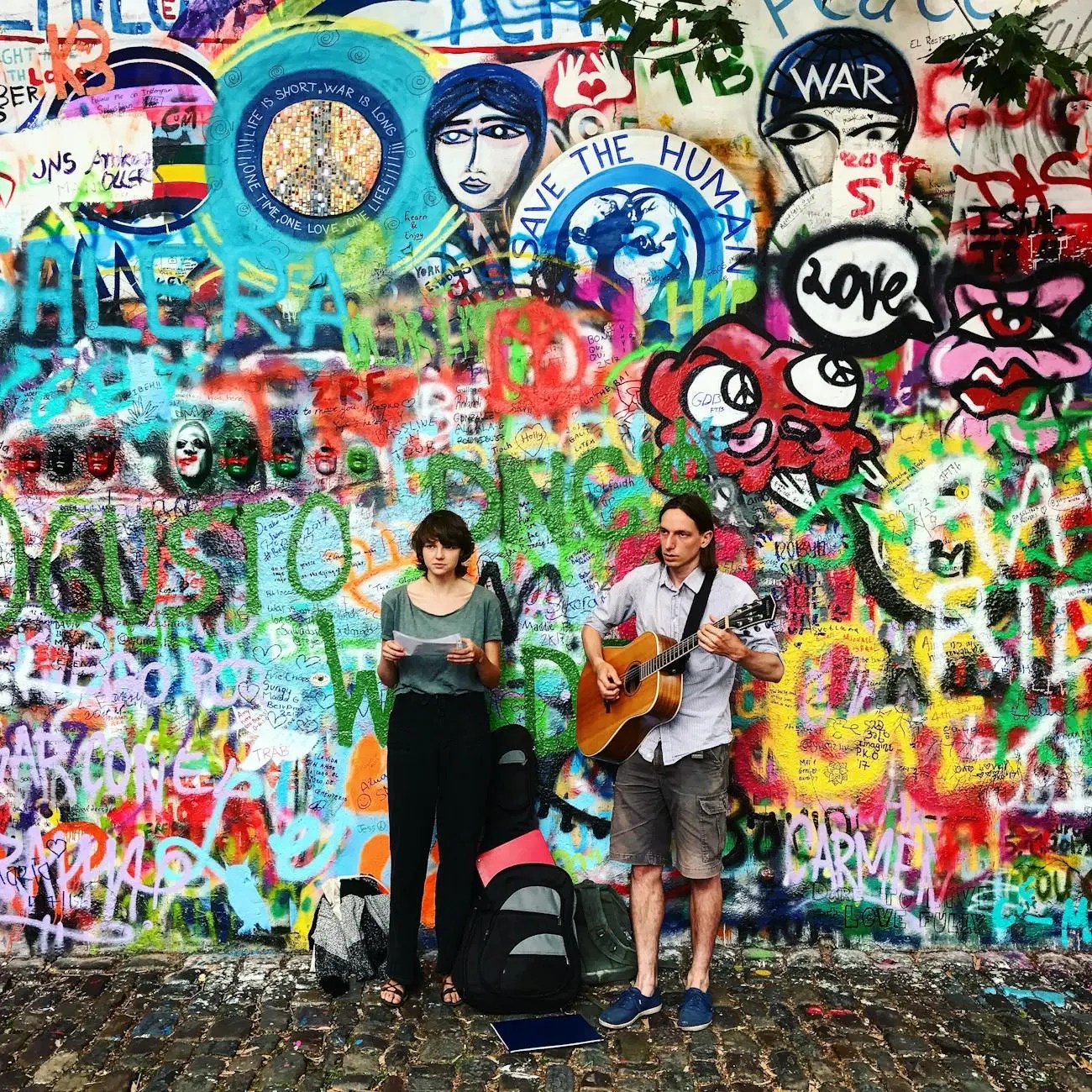 A man playing guitar and a woman reading lyrics stand in front of the colorful John Lennon Wall, covered in graffiti and artistic messages.