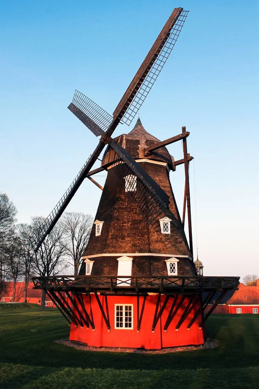A picturesque red windmill with wooden blades set against a clear blue sky, surrounded by green grass and trees.
