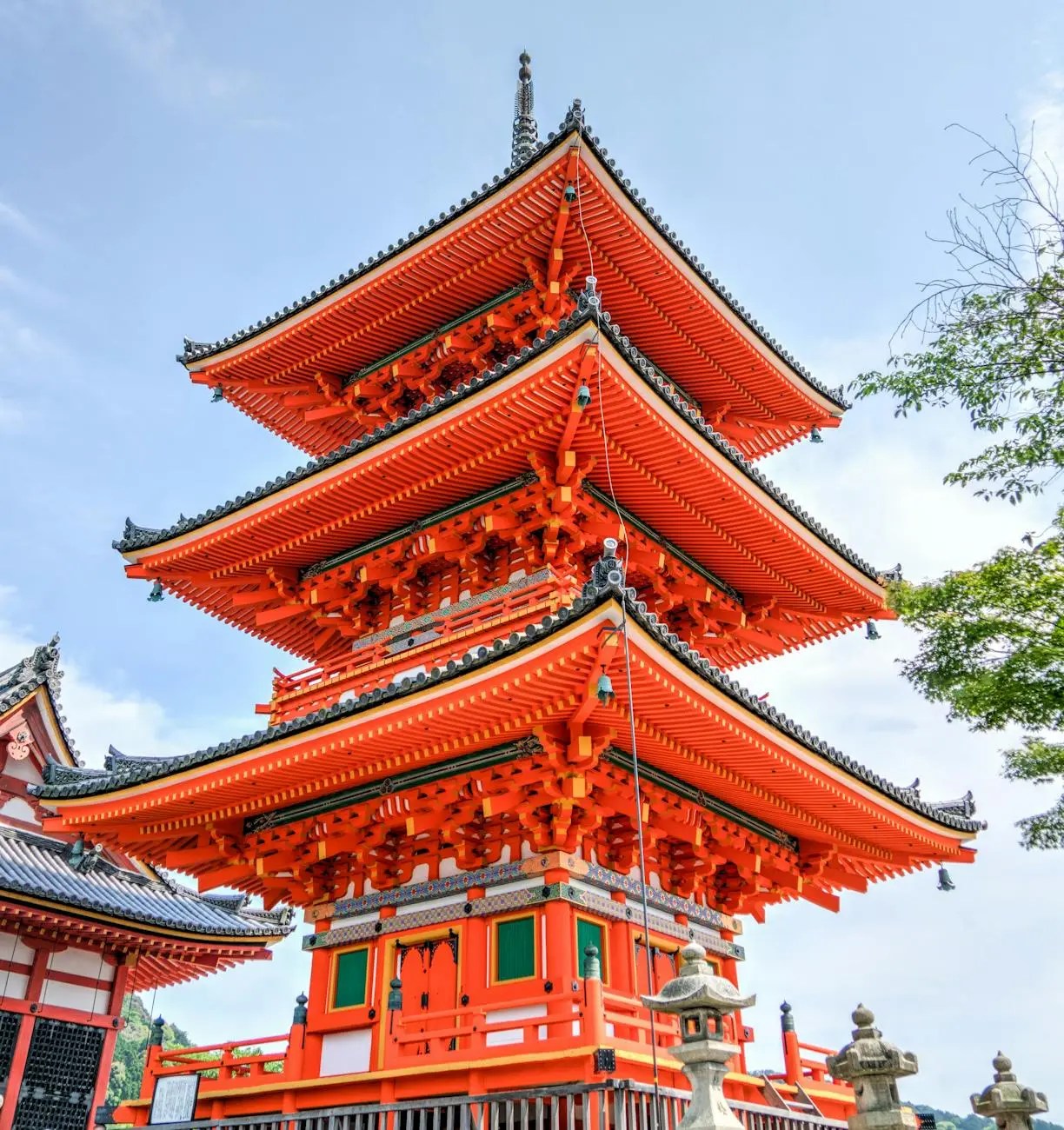 A vibrant three-story pagoda with intricate wooden details and a striking orange color, surrounded by lush greenery and a clear blue sky in Kyoto, Japan.