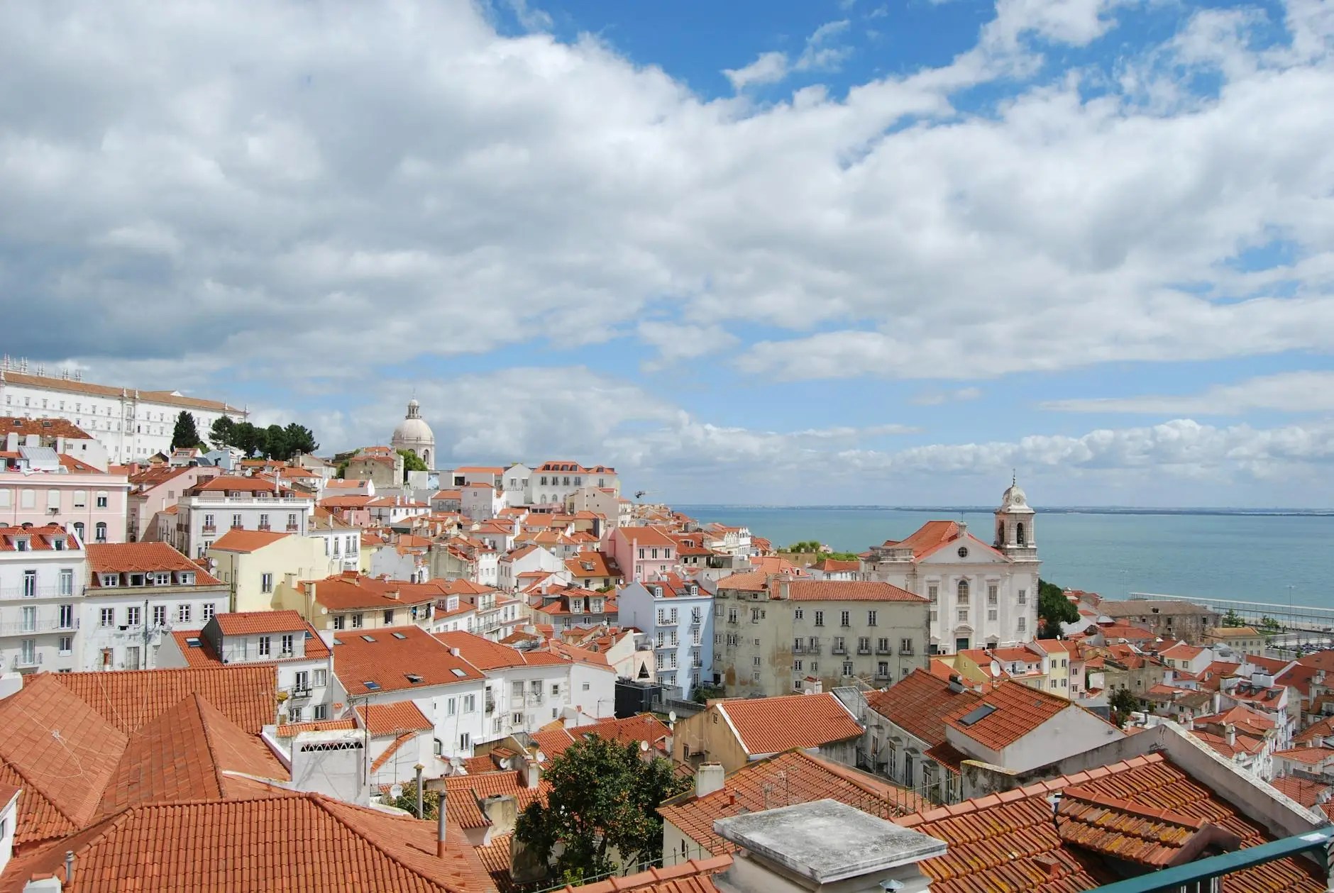 Panoramic view of Lisbon featuring terracotta rooftops, historical buildings, and distant views of the river under a partly cloudy sky.