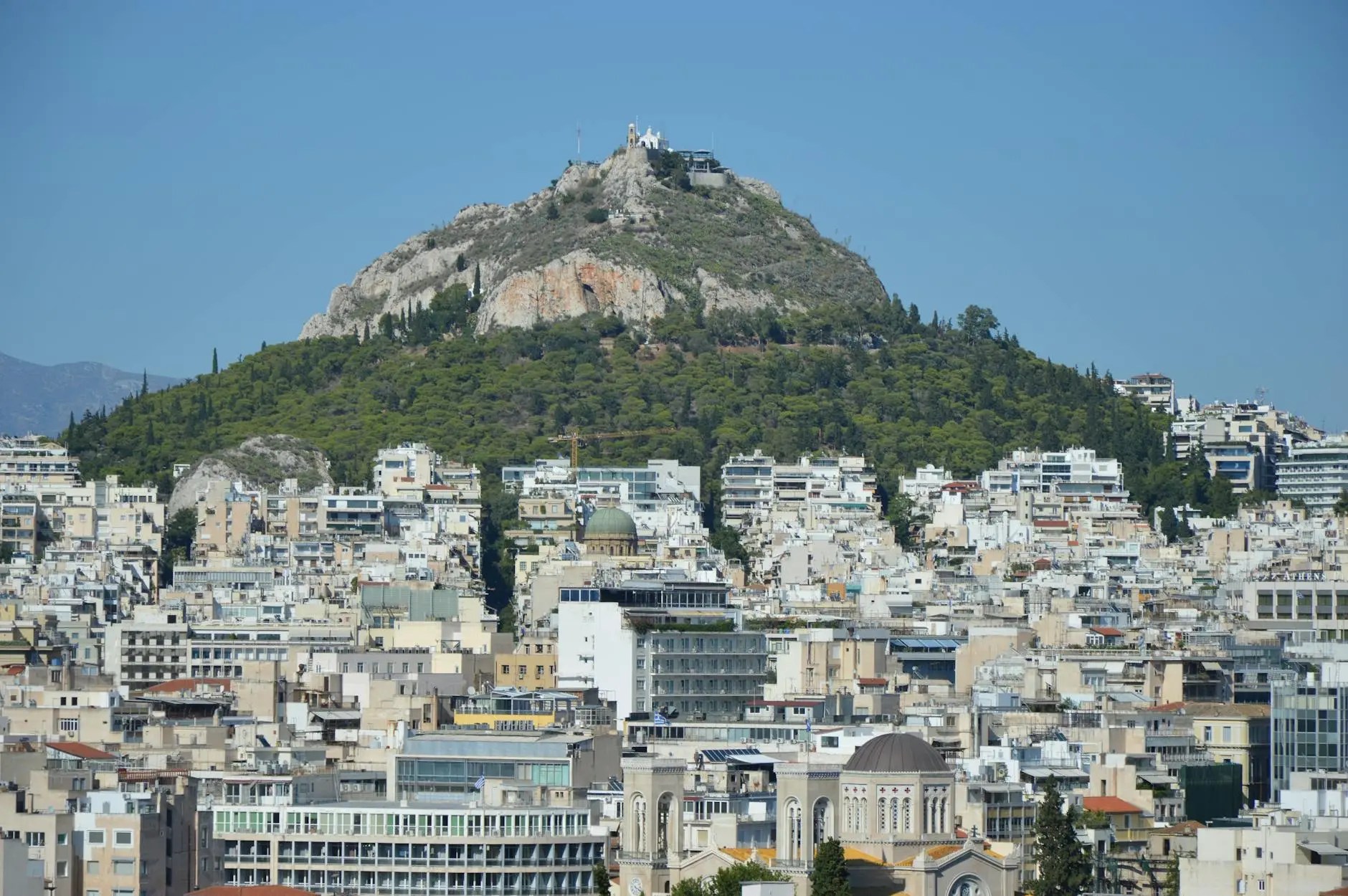 A panoramic view of Athens showcasing the dense urban landscape with a prominent green hill in the background, likely Lycabettus Hill, under a clear blue sky.