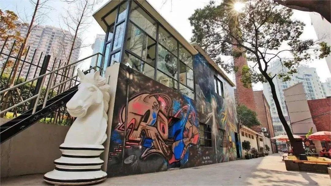 Exterior view of an art studio featuring a large white horse sculpture and vibrant graffiti on the wall, surrounded by trees and modern buildings.